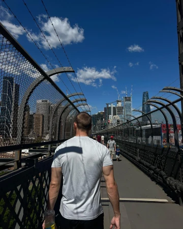 Man walking away on a fenced pedestrian walkway with city skyline and blue sky in Sydney, by Lachie Brycki