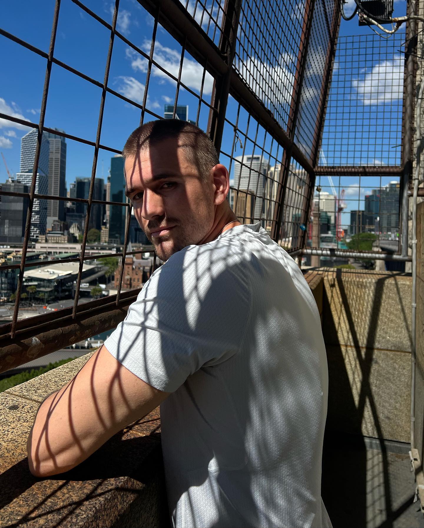 Man stands on a building looking at city skyline behind a steel cage. The bright daylight shot emphasizes the urban environment.