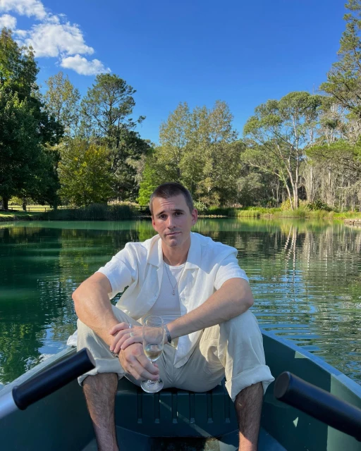 Adult male seated in a boat on a lake surrounded by trees on a sunny day, holding two champagne flutes with blue sky.