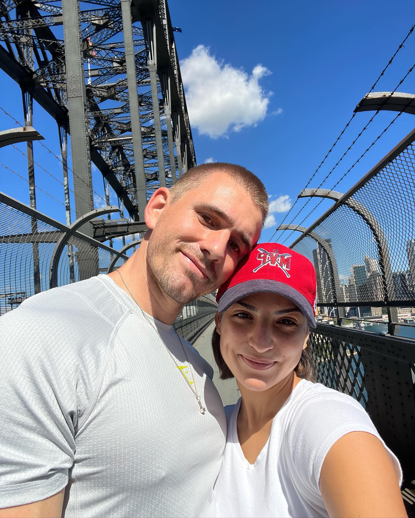 Couple smiles on Sydney Harbour Bridge with cityscape backdrop on a sunny day. Clear blue sky and bridge framework.