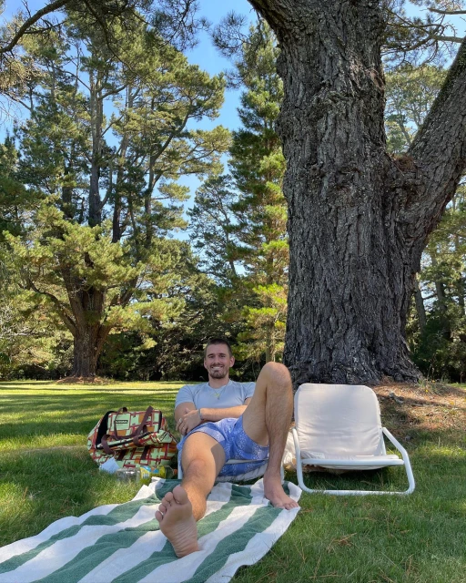 Adult male relaxing on striped towel in grassy area near large tree with a beach chair and bag in the background on a sunny day.