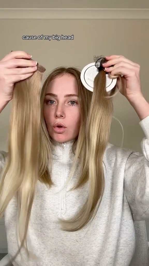 A blonde woman holds up two sets of long blonde hair extensions against a neutral background in a medium shot.