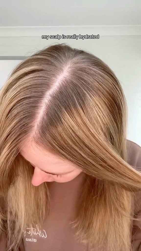 Close-up of a woman's blonde hair and scalp, showcasing her hair parting and hair texture, indoors with white wall background.