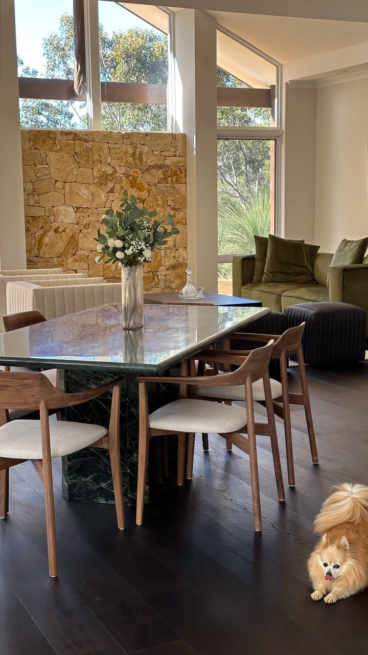 Interior view of dining room with stone wall, large windows, dining table, wood chairs, and dog in a home with dark wood floors.