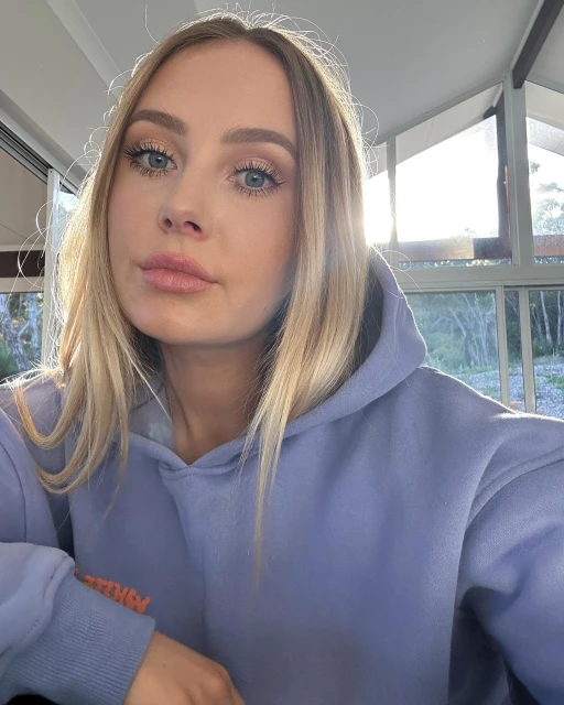 Blonde woman in a periwinkle hoodie poses indoors near windows with natural light. Focus on the human face and upper torso.