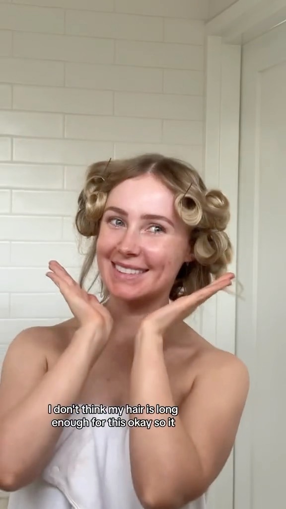 Smiling woman with fair skin and blonde hair wrapped in hair curlers with towel wrapped around her body, against a white tiled bathroom.