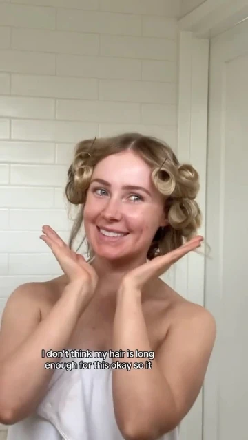 Smiling woman with fair skin and blonde hair wrapped in hair curlers with towel wrapped around her body, against a white tiled bathroom.