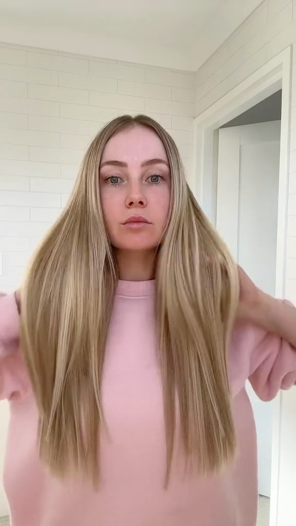 Woman with long blonde hair wearing a pink sweatshirt in an indoor setting with a white tiled wall and doorway.