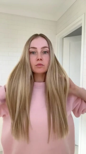 Woman with long blonde hair wearing a pink sweatshirt in an indoor setting with a white tiled wall and doorway.