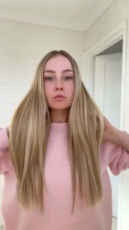 Woman with long blonde hair wearing a pink sweatshirt in an indoor setting with a white tiled wall and doorway.