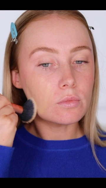 Blonde woman applying foundation with a brush in a close-up shot, showcasing makeup application and beauty routine in a studio setting.