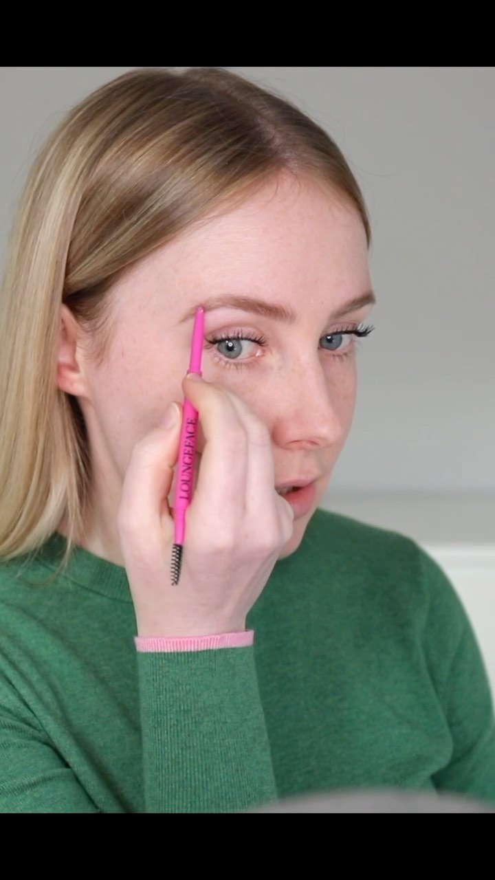 Close-up of a fair-skinned blonde woman applying makeup to her eyebrow with a pink eyebrow brush with a grey background and green shirt.