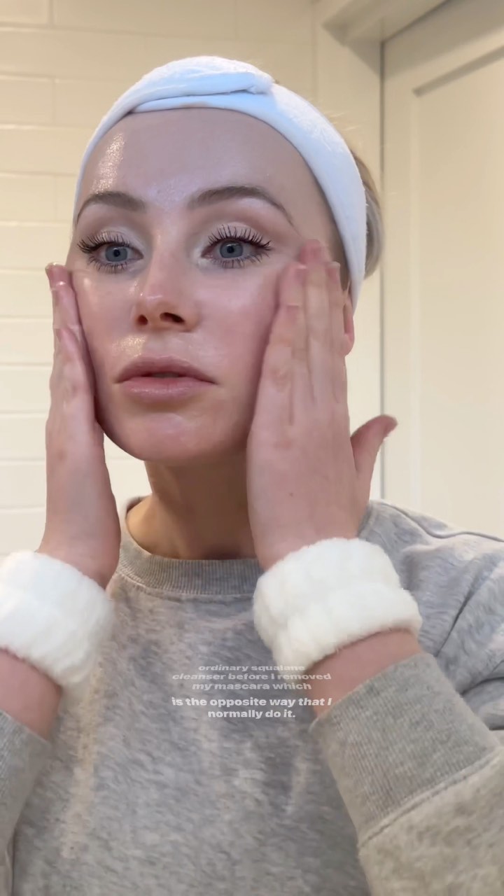 Adult woman with blond hair wearing a white headband and wristbands, applying skincare to her face in a bathroom setting.