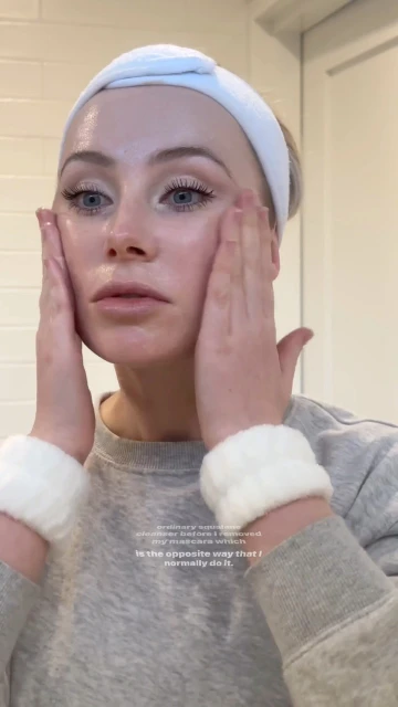Adult woman with blond hair wearing a white headband and wristbands, applying skincare to her face in a bathroom setting.