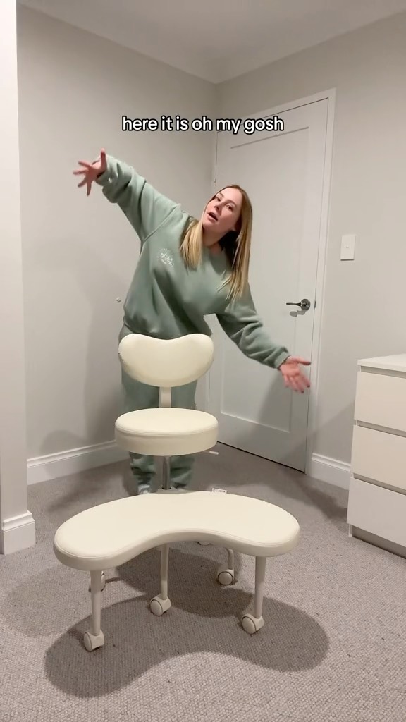 Woman stands beside ergonomic kneeling chair in an indoor setting. The chair is cream-colored, against neutral walls and carpet.