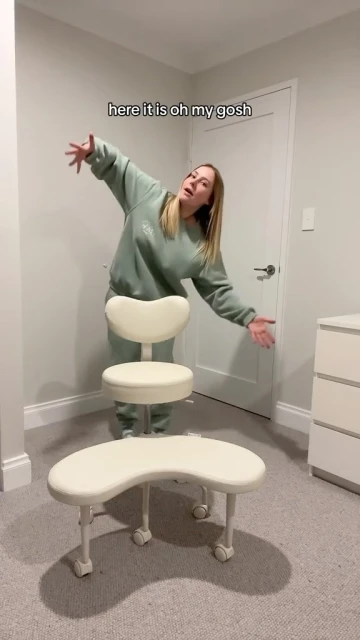 Woman stands beside ergonomic kneeling chair in an indoor setting. The chair is cream-colored, against neutral walls and carpet.
