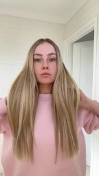 Blonde woman wearing a pink sweatshirt holds up her long straight hair in a bright room with white tile walls.