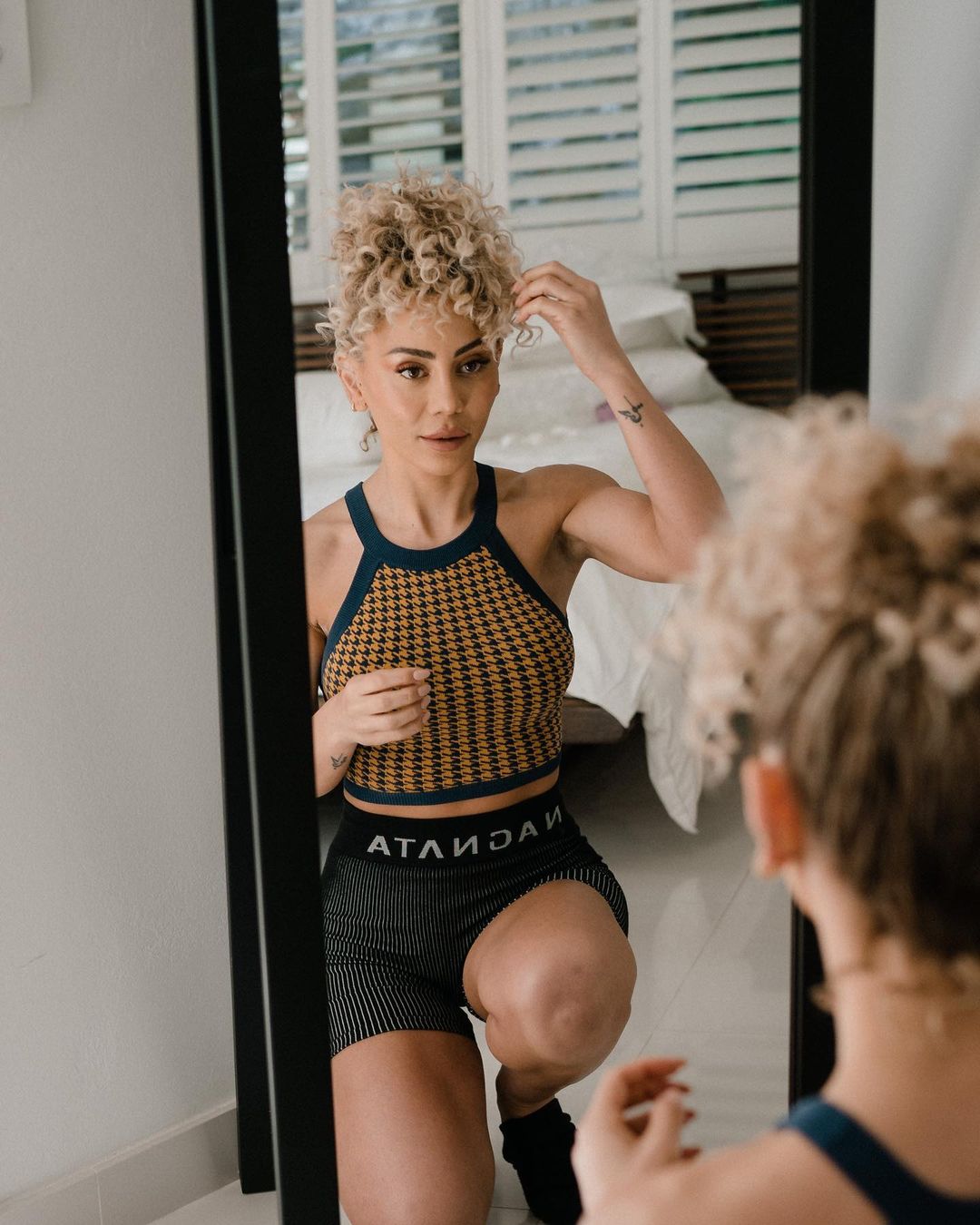 Athletic woman with curly hair kneels facing a mirror in her home, wearing a knit halter top and striped shorts, showcasing fitness.