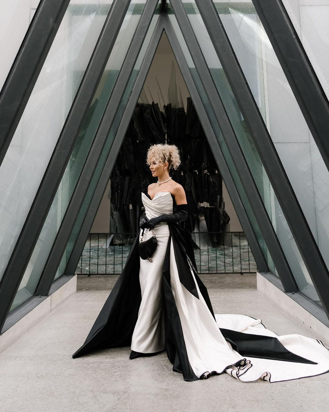 Elegant woman with blonde hair poses in a black and white gown with matching opera gloves and small purse against a modern building.