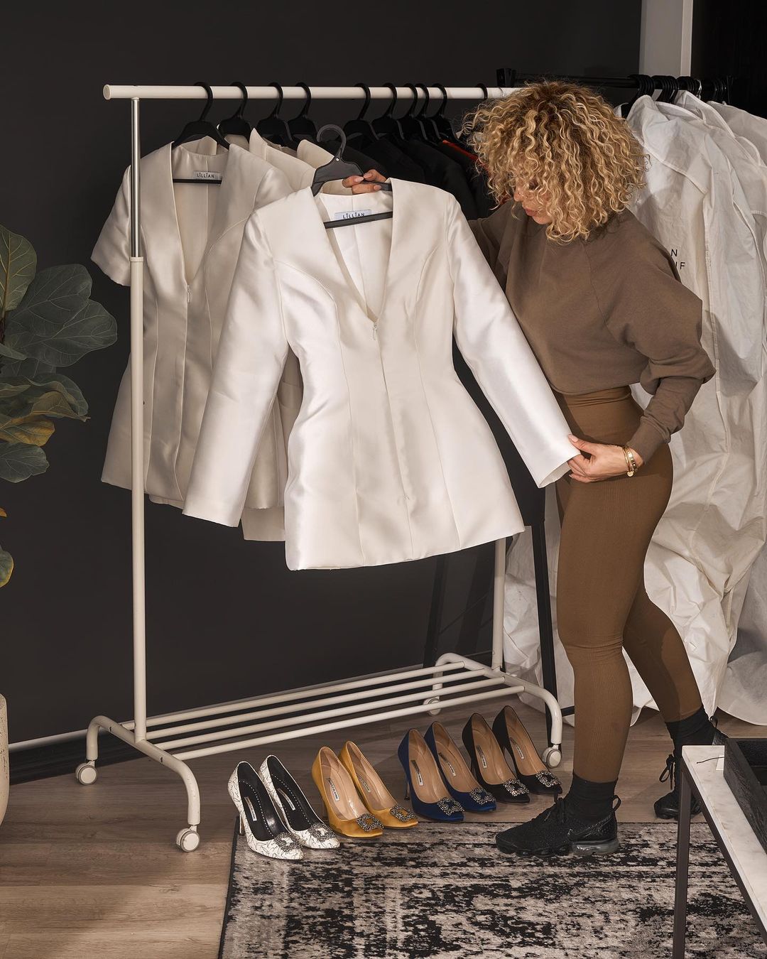 Woman with curly hair examines a white jacket on a rack with shoes beneath, in a fashion-forward indoor setting, against a dark wall.