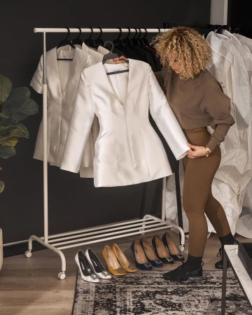 Woman with curly hair examines a white jacket on a rack with shoes beneath, in a fashion-forward indoor setting, against a dark wall.