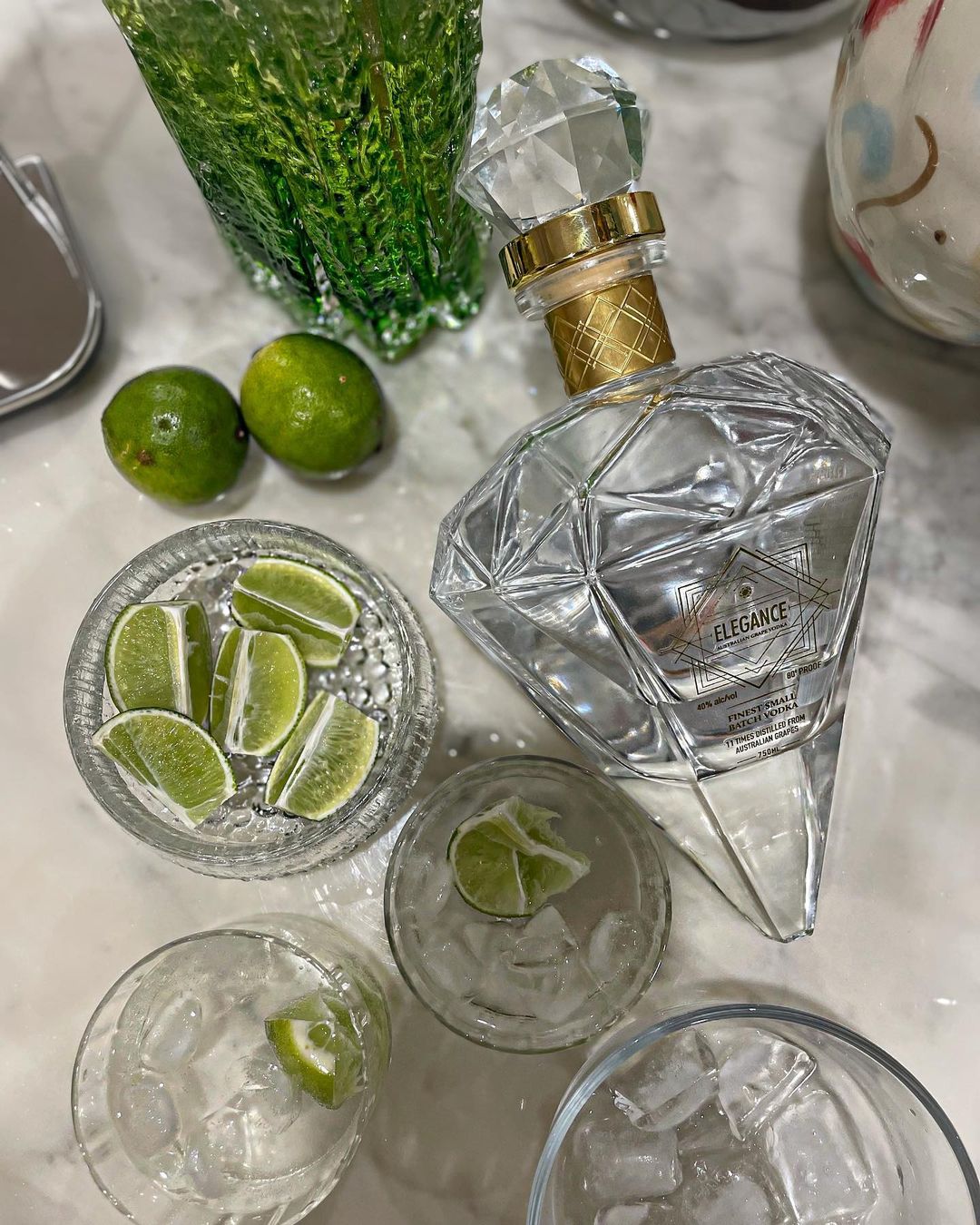 Overhead shot of a diamond-shaped liquor bottle surrounded by limes, cut limes in a bowl, and glasses with ice, on a white surface.