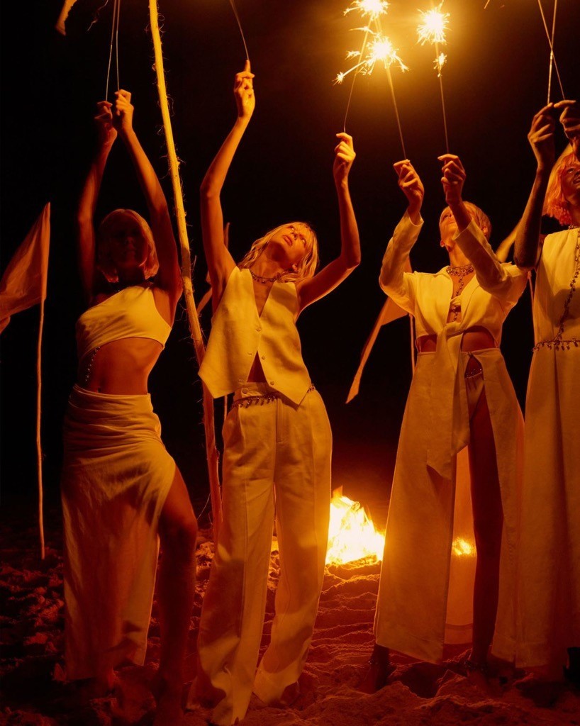 Group of young adult women in white clothing stand outside at night, holding sparklers around a fire in an outdoor setting.