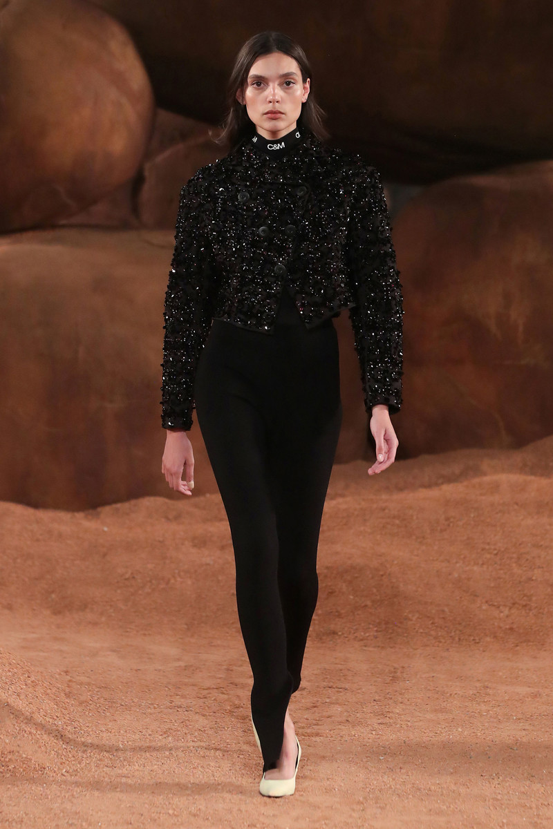 Runway fashion: a slender woman in a black beaded jacket and leggings walks the runway, a neutral, earthen-toned backdrop completing the scene.