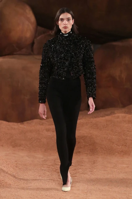 Runway fashion: a slender woman in a black beaded jacket and leggings walks the runway, a neutral, earthen-toned backdrop completing the scene.