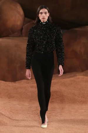 Runway fashion: a slender woman in a black beaded jacket and leggings walks the runway, a neutral, earthen-toned backdrop completing the scene.