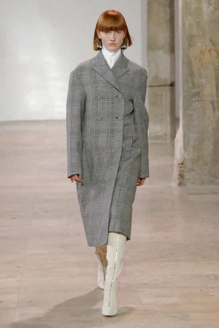 Fashion model walks the runway wearing a grey plaid coat and white boots in an indoor studio with neutral background during a show.