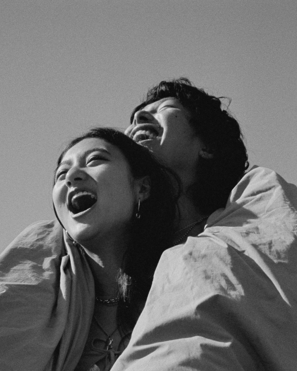 A happy couple laughs, embracing beneath a clear sky. The black and white photo captures their joyful moment in natural light.