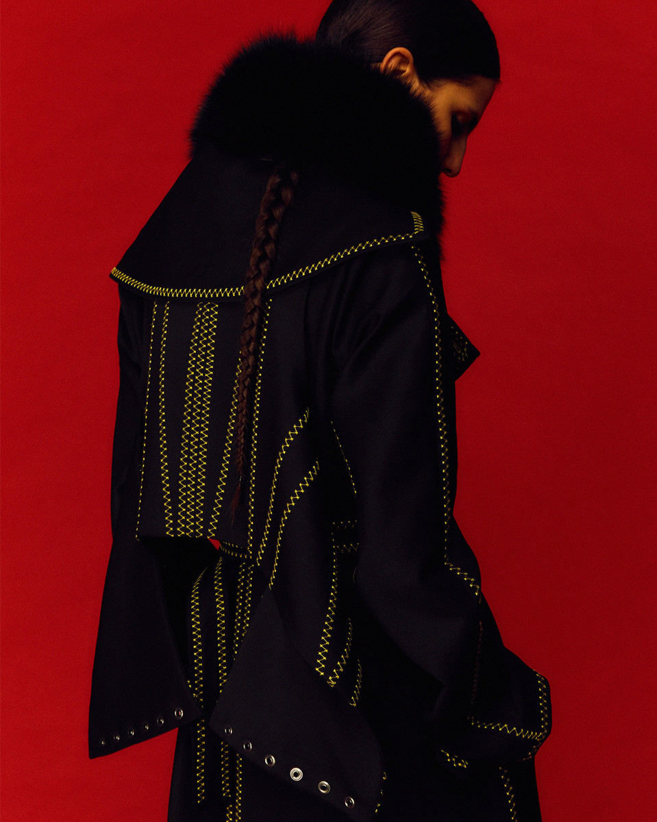Fashion shot: person in a black coat with fur collar and yellow stitching on red background. Braided hair is visible.