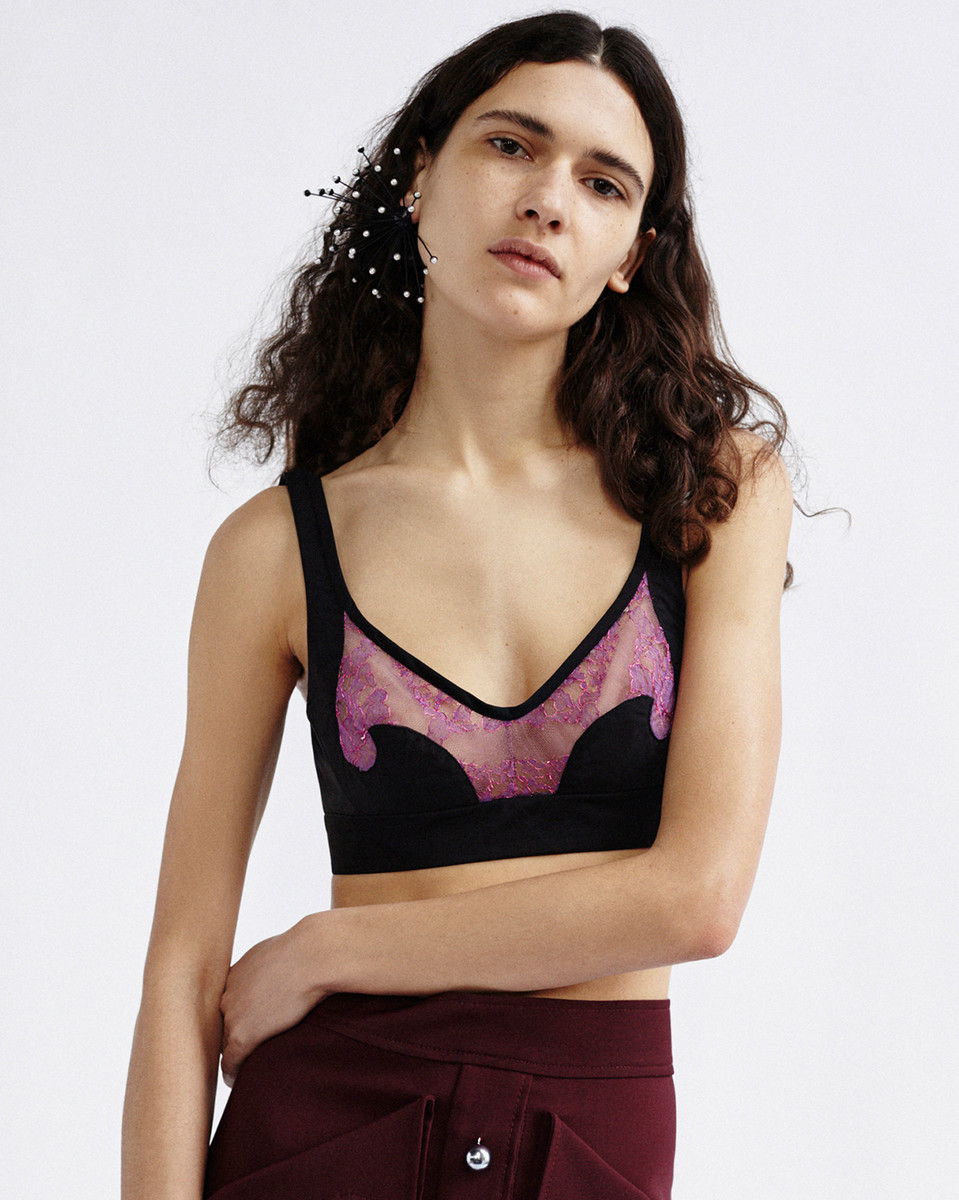 A young adult female model poses indoors, wearing a black and pink bra top and a maroon skirt against a white backdrop.