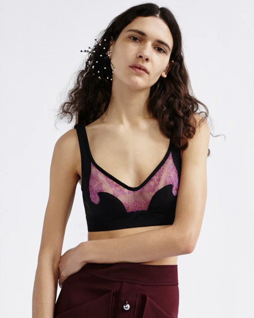 A young adult female model poses indoors, wearing a black and pink bra top and a maroon skirt against a white backdrop.