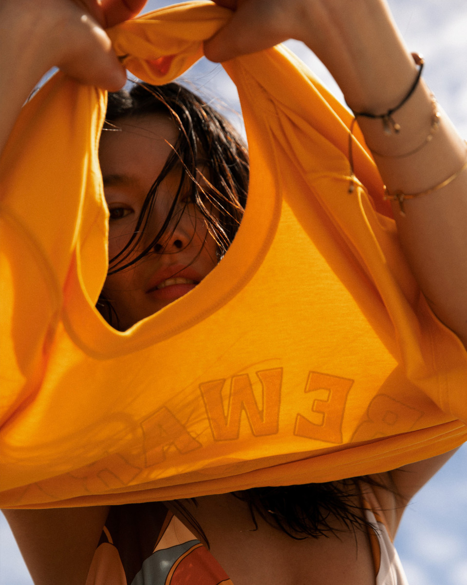 Woman holding up a bright yellow garment with raised lettering, peering through the neckline against a sky background.
