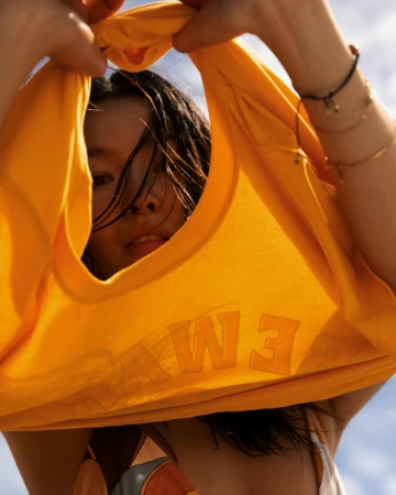 Woman holding up a bright yellow garment with raised lettering, peering through the neckline against a sky background.