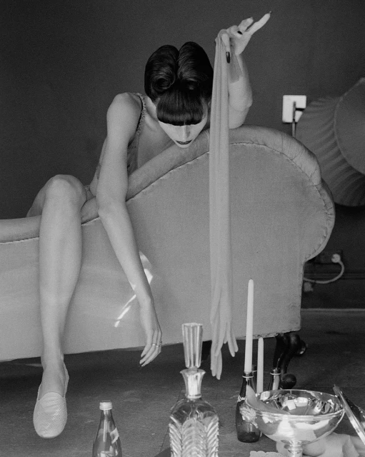 Black and white portrait of a woman reclining on a chaise lounge holding hosiery, next to candles and glass bottles.