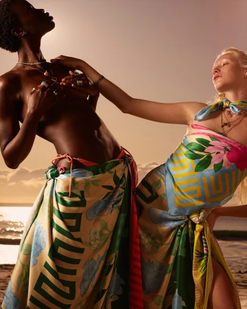 Two diverse models in patterned wraps pose on a beach at sunset. The shot focuses on their upper bodies with a warm color grading.