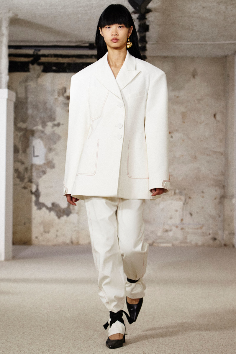 Model with dark hair wearing an oversized white blazer and matching trousers walking in an interior space