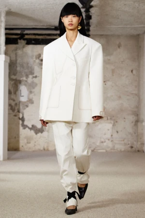 Model with dark hair wearing an oversized white blazer and matching trousers walking in an interior space