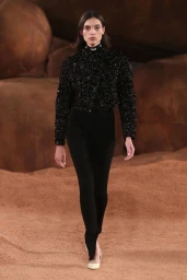 Model walking a runway wearing a black textured jacket and fitted trousers against a rocky earth backdrop