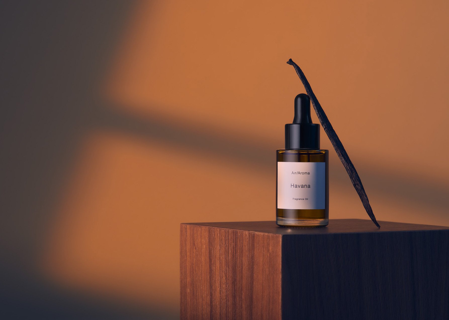 Close-up of a brown glass dropper bottle with vanilla bean on a wooden cube against an orange background with light and shadow play.