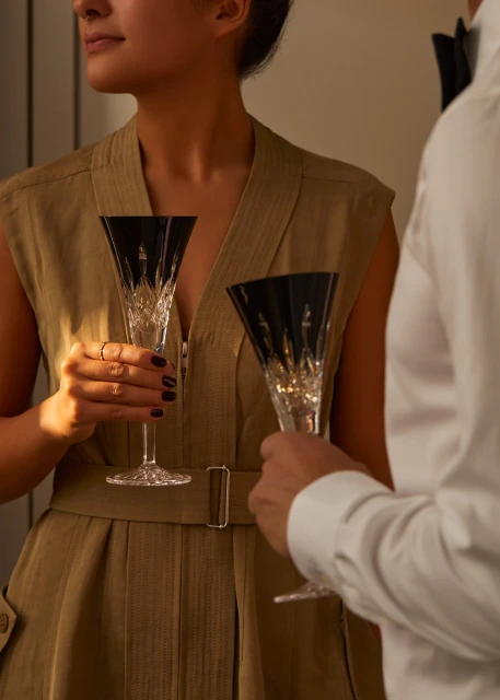 Close-up of a partially visible couple holding champagne flutes with black tops while wearing a khaki dress and white shirt.