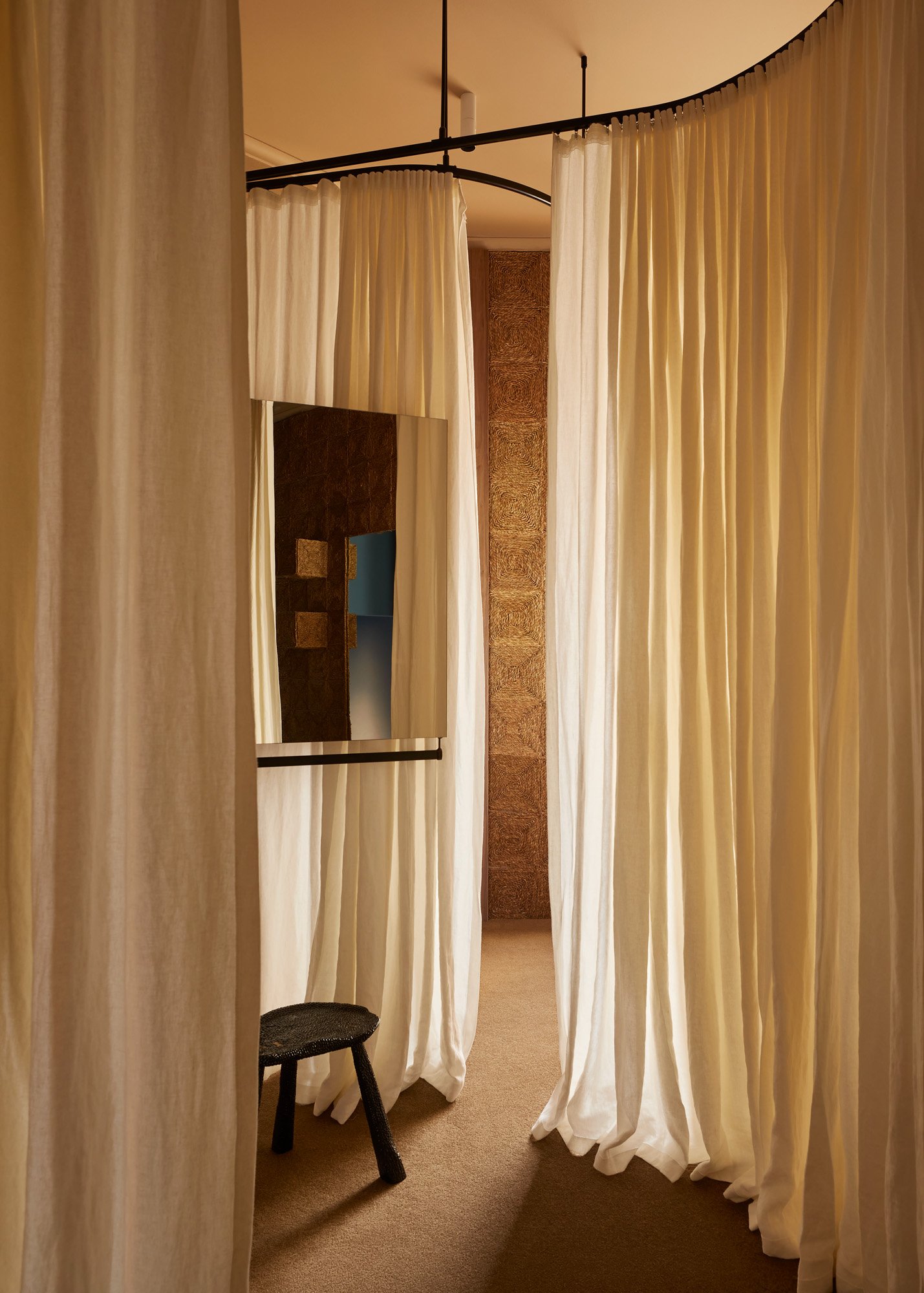 Interior view of a room featuring draped curtains and a small, dark wood stool, illuminated by soft lighting, creating a cozy atmosphere.