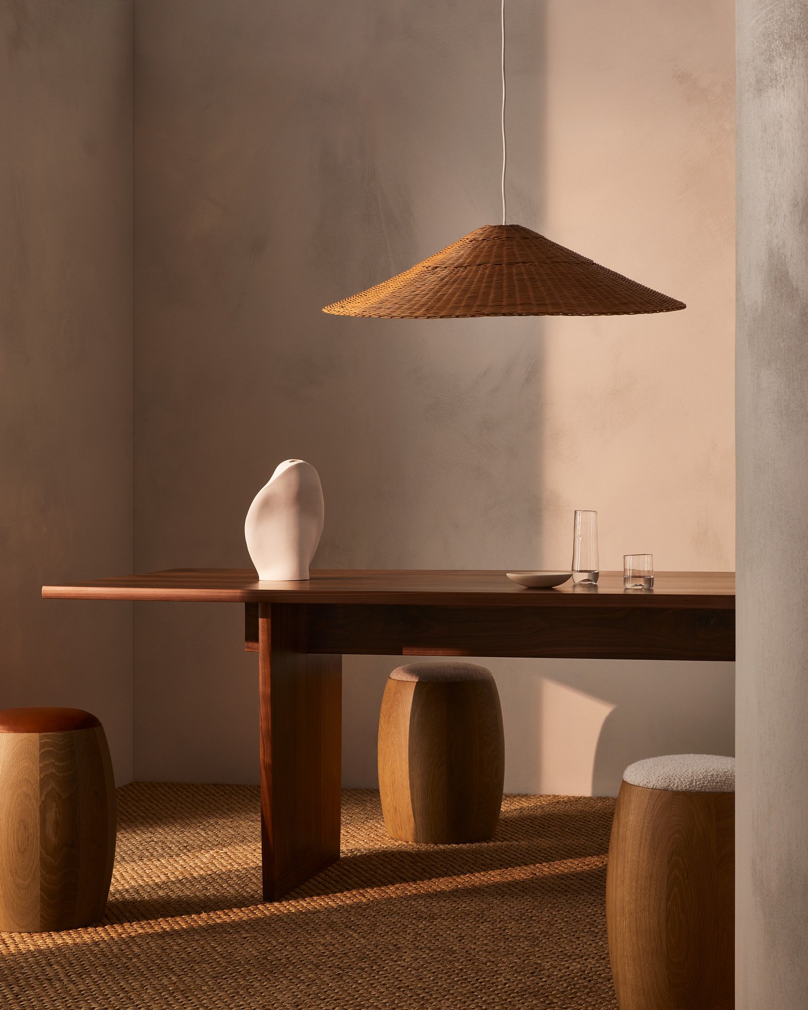 Minimalist interior featuring a wood dining table, woven pendant light, ceramic vase, wood stools, and neutral color scheme creating a warm ambiance.