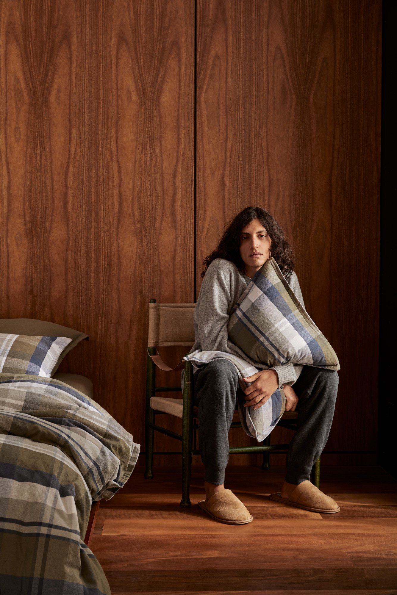 Adult male with long hair sits on a chair and hugs a plaid pillow. The room has wooden paneled walls and a bed with plaid bedding.