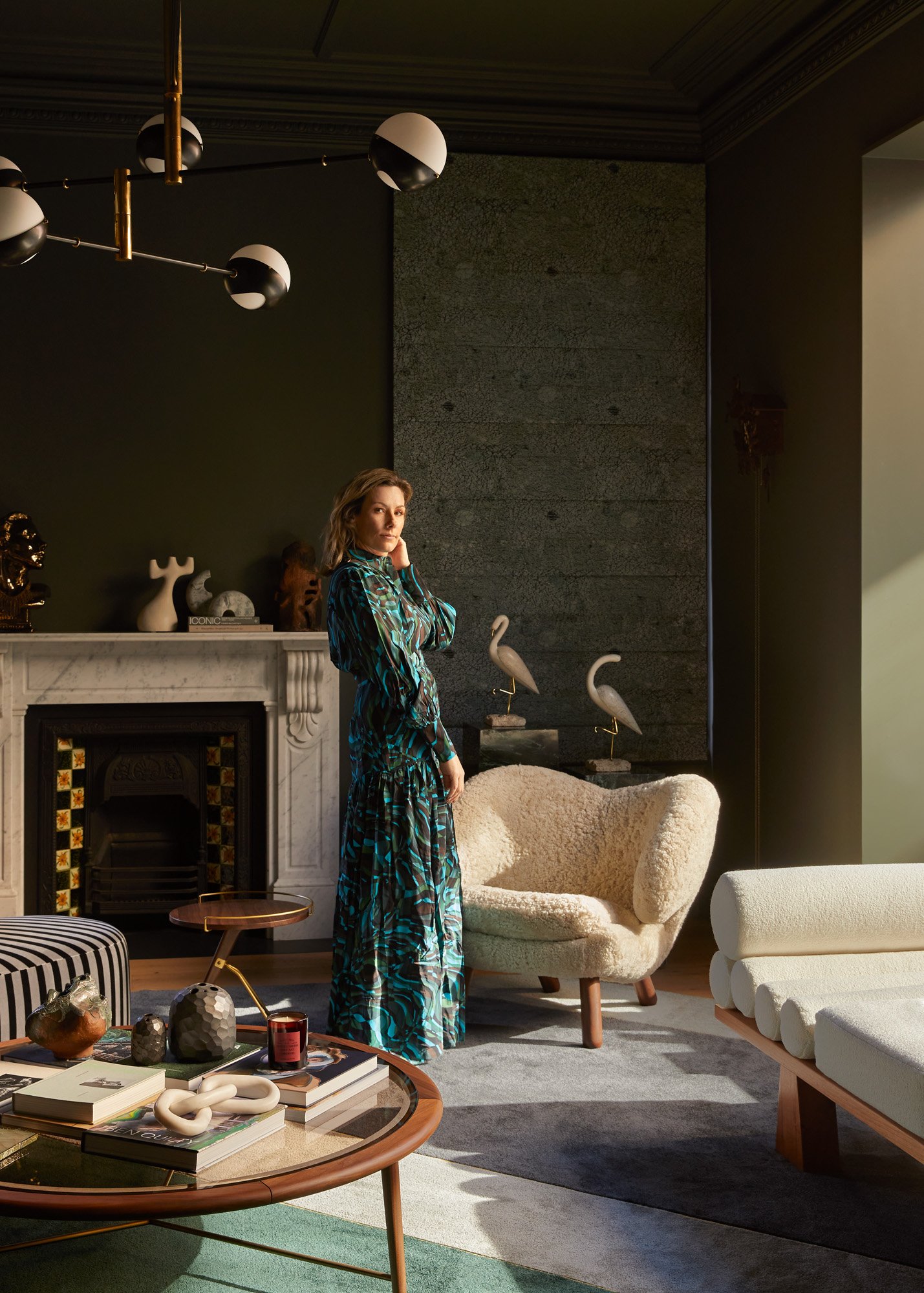 Woman in a patterned dress stands in a living room with modern decor, including a fireplace, unique furniture, and a contemporary lighting fixture.
