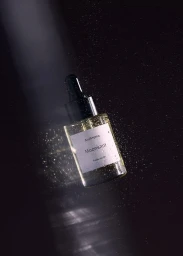 A glass dropper bottle of perfume oil floating against a dark textured background with water droplets and a bright vertical light beam.