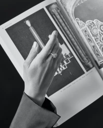 Close-up black and white photograph of a woman's hand wearing a ring against an open book with architectural imagery.